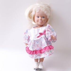 May include: A vintage doll with blonde hair and blue eyes, wearing a white dress with a pink bow and floral print. The dress has pink ruffles at the hem. The doll is wearing white roller skates.