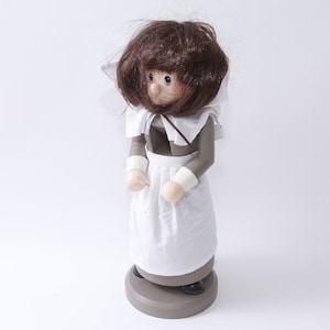 May include: A wooden figurine of a pilgrim woman wearing a brown dress, white apron, and white collar. She has brown hair and is standing on a brown base.