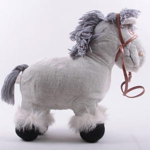 May include: A plush toy horse in a standing position. The horse is primarily light gray with a gray mane and tail. It has white fur around its hooves and black hooves. A brown bridle is around the horse's head.