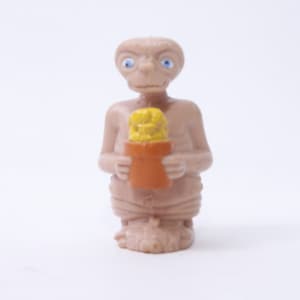 Puede incluir: Una pequeña figura de plástico marrón de un extraterrestre sosteniendo una caja amarilla y naranja. El extraterrestre tiene ojos grandes y azules y una boca ancha.