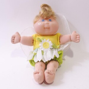May include: A vintage doll with blonde hair styled in a swirl. The doll wears a yellow ribbed top, a green and white floral skirt, and has blue eyes. It has a daisy on the front and white wings.