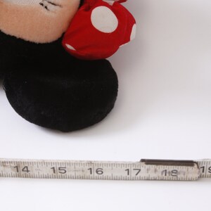 May include: A close-up of a black and red plush toy mouse with white polka dots on the red bow. The toy is lying on a white surface with a ruler next to it.