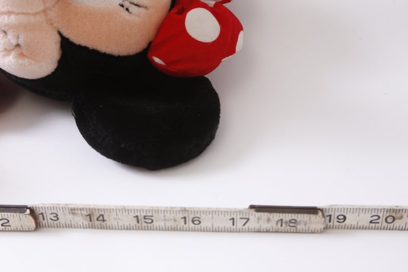 May include: A close-up of a black and red plush toy mouse with white polka dots on the red bow. The toy is lying on a white surface with a ruler next to it.