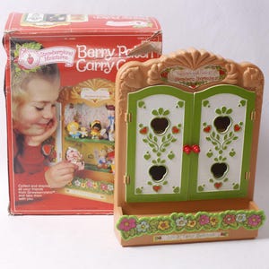 May include: A tan plastic Strawberryland Miniatures Berry Patch Carry Case with green doors featuring strawberry-shaped cutouts and red knobs. The base is decorated with colorful painted flowers. The original red cardboard box is included.