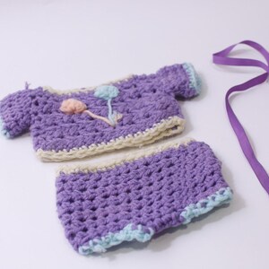 May include: A handmade crochet outfit in shades of purple, cream, and light blue. The set includes a short-sleeved top and matching shorts, with floral embellishments. A purple ribbon lies nearby.
