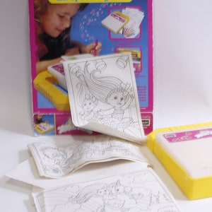 May include: A yellow and pink Magic Trolls Lite-Up Drawing Desk with a light-up drawing surface and a set of colorful markers. The box includes several drawing sheets with images of trolls and unicorns.