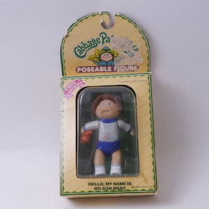 Peut inclure: Une figurine articulée Cabbage Patch Kids vêtue d'une tenue bleu et blanche tenant un ours en peluche. La figurine est dans une vitrine en plastique avec une boîte en carton jaune et verte. La boîte indique "Première édition" et "Figurines articulées". Le texte sur la boîte indique également "Bonjour, je m'appelle Wilson Marc".