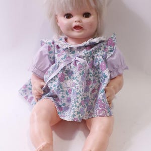 May include: A vintage baby doll with blonde hair and brown eyes, wearing a floral print dress with purple sleeves. The doll is sitting with its legs extended, and has a sweet expression.