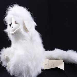 May include: A plush toy monkey covered in long, white faux fur with a smooth, light-colored face, dark brown eyes, and a long, furry tail. A small, rectangular paper tag with handwritten text is attached to the base of the toy's body.