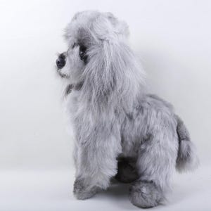 May include: A plush, gray poodle toy with a fluffy, textured coat. The toy dog is posed in a seated position, with its head turned to the left. The poodle's fur is a soft, light gray color, and its features are detailed.