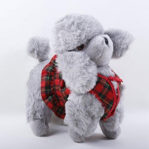 May include: A plush toy poodle with soft grey fur, wearing a red and black plaid dress with a ruffled skirt. The stuffed animal has dark eyes, a black nose, and a pom-pom tail, standing in a classic pose against a plain white background.