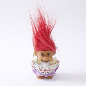 May include: A red-haired troll doll ornament wearing a purple dress and holding a banner that says "Feliz Navidad".