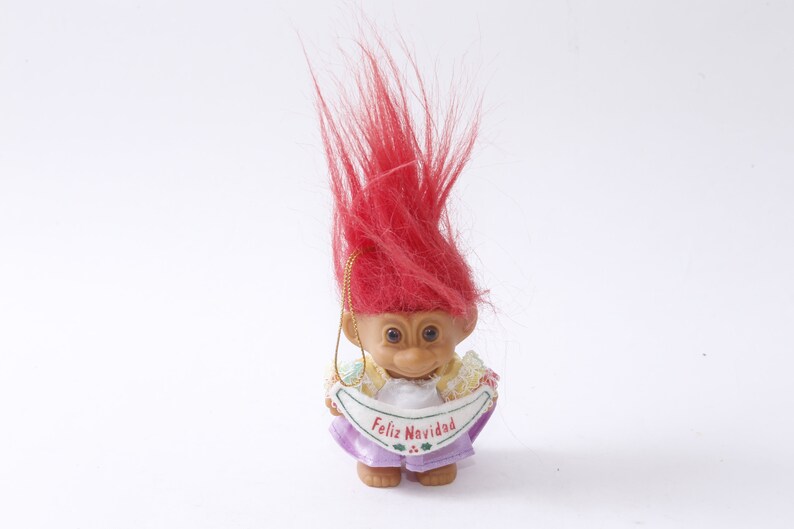 May include: A red-haired troll doll ornament wearing a purple dress and holding a banner that says "Feliz Navidad".