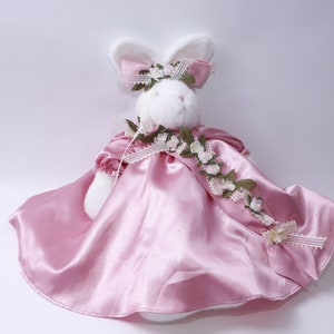 Peut inclure: Un lapin en peluche blanc portant une robe en satin rose avec une guirlande florale et un n&oelig;ud blanc.