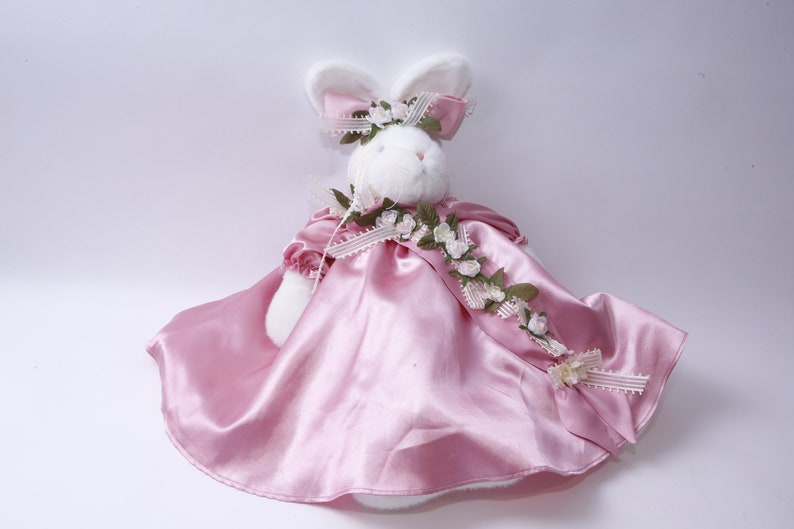 Peut inclure: Un lapin en peluche blanc portant une robe en satin rose avec une guirlande florale et un n&oelig;ud blanc.