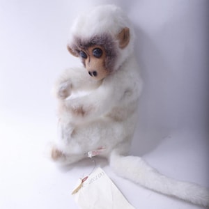 May include: A plush white monkey with a brown face and ears, and blue eyes. The monkey is sitting with its arms crossed and a long tail. A tag is attached to the monkey.