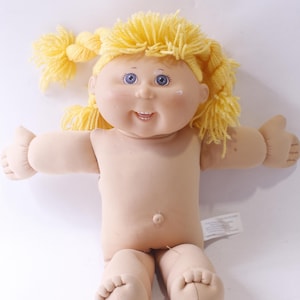 May include: A Cabbage Patch Kid doll with blonde yarn hair, blue eyes, and a smiling face. The doll is sitting with its legs crossed and is wearing no clothes.