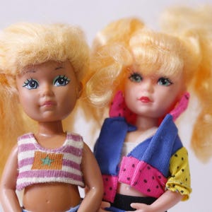 May include: Two vintage dolls with blonde hair and blue eyes. One doll wears a striped crop top and the other a colourful jacket with pink polka dots. The dolls are a nostalgic representation of 1980s fashion.