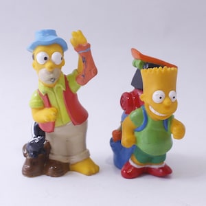 May include: Two yellow plastic Simpsons figures. The figure on the left is wearing a blue hat, a red vest, and holding a black and white skunk. The figure on the right is wearing a green shirt and a blue backpack with a skateboard on top.