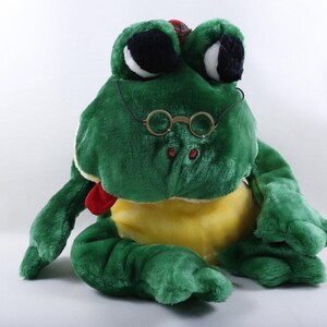 May include: A plush frog toy with bright green fur, a yellow belly, and large black and white eyes. The frog wears thin, round, gold-toned wire-rimmed glasses and has a small red tongue protruding from its mouth. The toy is seated in a forward-facing position.