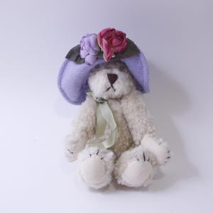 May include: A small, white teddy bear wearing a lavender hat with pink and purple flowers. The bear is sitting on a white background.