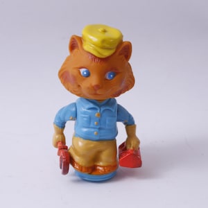 May include: A toy figurine of a cat wearing a yellow hat and blue overalls. The cat is holding a wrench in one hand and a red toolbox in the other.