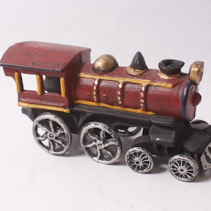Op de afbeelding: Een decoratief modeltrein van gietijzer, geschilderd in tinten rood, zwart en zilver. De trein heeft een gedetailleerd ontwerp met zichtbare wielen, een schoorsteen en een cabine. Het model is ongeveer 15 cm lang.