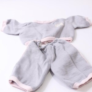 May include: A grey doll's outfit, comprising a short-sleeved top and matching shorts. The edges of the top and shorts are trimmed with pink fabric. A small white design is on the top.
