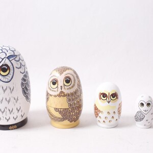 May include: A set of five wooden nesting dolls, each painted to resemble an owl. The largest doll is white with blue and black details, while the others feature varying colors and patterns, including brown, yellow, and orange.