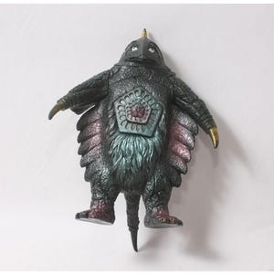 May include: A dark, textured monster figurine with a pointed head, gold-tipped claws, and a prominent pentagonal chest emblem. The creature features a mix of black, metallic blue, and reddish-purple hues, with a thick, fur-like texture on its torso.