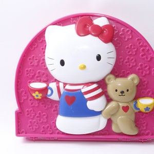 May include: A pink plastic Hello Kitty themed lunchbox with a 3D image of Hello Kitty and a teddy bear holding teacups. The lunchbox has a flower pattern.