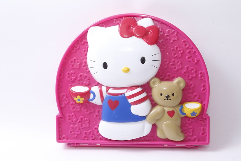 May include: A pink plastic Hello Kitty themed lunchbox with a 3D image of Hello Kitty and a teddy bear holding teacups. The lunchbox has a flower pattern.