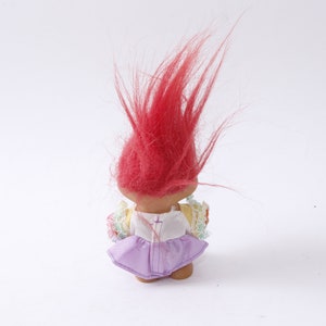 May include: A red-haired troll doll wearing a white dress with a purple skirt and floral accents.