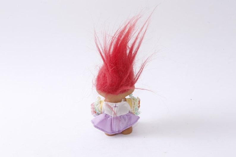 May include: A red-haired troll doll wearing a white dress with a purple skirt and floral accents.