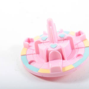 May include: A pink plastic carousel with four seats and a central pole. The carousel has a blue and yellow striped base.