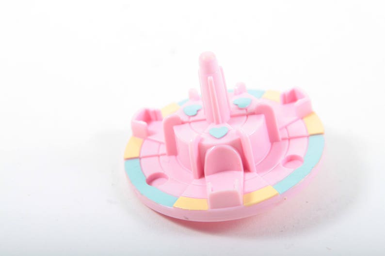 May include: A pink plastic carousel with four seats and a central pole. The carousel has a blue and yellow striped base.