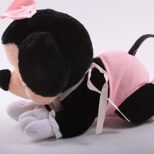 May include: A plush toy of a black and white mouse wearing a pink dress and a pink bow. The mouse is lying on its belly with its head turned to the side.