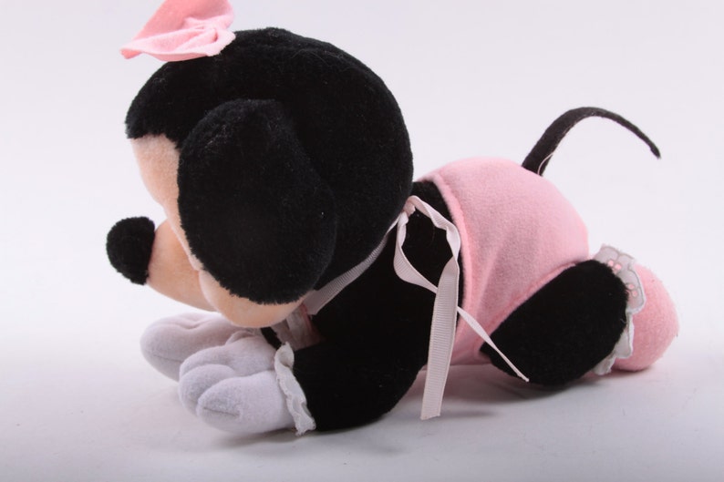 May include: A plush toy of a black and white mouse wearing a pink dress and a pink bow. The mouse is lying on its belly with its head turned to the side.