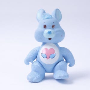May include: A light blue Care Bear-like figurine with a rabbit-like appearance. It has a pink heart with wings on its chest and small red accents on its face. The toy is made of plastic and has a soft, plush-like texture.