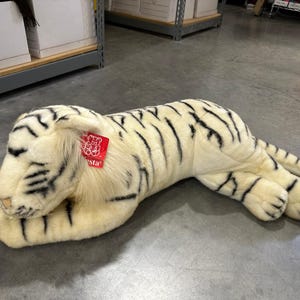 Pode incluir: Um grande tigre branco de pelúcia com listras pretas, deitado em um chão de concreto. O bicho de pelúcia tem uma etiqueta vermelha presa à orelha. O tigre é um brinquedo macio, perfeito para crianças ou colecionadores.