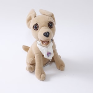May include: A tan plush dog toy with large, expressive eyes and floppy ears. The dog is wearing a white bandana with a purple logo. The toy is sitting upright on a white background.