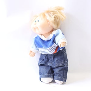 Puede incluir: Una muñeca vintage Cabbage Patch Kid con cabello rubio, ojos azules y una camisa a rayas azules y blancas. La muñeca lleva pantalones vaqueros y está de pie sobre un fondo blanco.