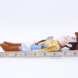 Może przedstawiać: Pluszowa zabawka Woody'ego z serii Toy Story. Woody leży na boku z wyciągniętą prawą ręką. Ma na sobie sw&oacute;j charakterystyczny kowbojski kapelusz, kamizelkę i dżinsy. Zabawka ma około 18 cm długości.