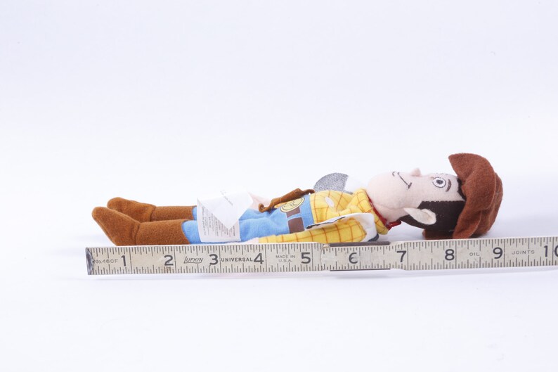 Może przedstawiać: Pluszowa zabawka Woody'ego z serii Toy Story. Woody leży na boku z wyciągniętą prawą ręką. Ma na sobie sw&oacute;j charakterystyczny kowbojski kapelusz, kamizelkę i dżinsy. Zabawka ma około 18 cm długości.
