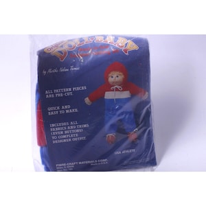 May include: A packaged doll-baby craft kit with a blue background. The package features a doll illustration wearing a red hooded jacket, white shirt, and blue pants. Text includes "Original DOLL-BABY" and "USA ATHLETE."