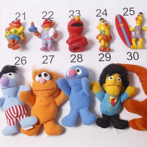 May include: A collection of Sesame Street character figurines, including Elmo, Bert, Ernie, and Grover. The figures are brightly colored and made of plastic or plush material. Each figure is numbered from 21 to 30.
