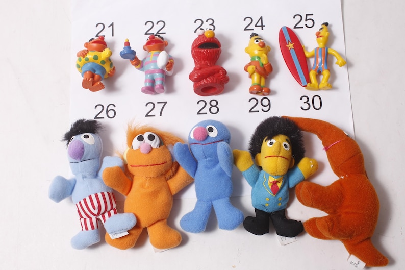 May include: A collection of Sesame Street character figurines, including Elmo, Bert, Ernie, and Grover. The figures are brightly colored and made of plastic or plush material. Each figure is numbered from 21 to 30.