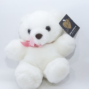 May include: White plush teddy bear with a pink bow, sitting on a white background. The bear has black eyes and a brown nose. The tag on the bear reads "With Love" and "Our Mill Enterprises Inc."