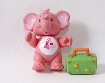 May include: A pink elephant toy with large ears and a heart emblem on its chest. Next to it is a green suitcase with an orange handle and yellow star decorations. A vintage toy.