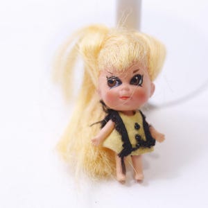 May include: A vintage doll with blonde hair styled in pigtails. The doll has a yellow dress with black trim and black buttons. The doll's face is painted with large eyes and rosy cheeks. The doll is standing on a white surface.